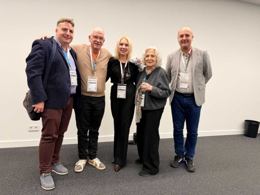 5 people with conference badge necklaces posed in front of a light colored wall 